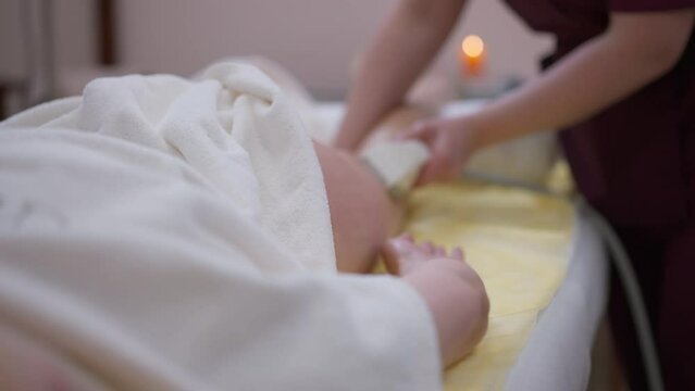 Close-up white towel with blurred masseur massaging client thigh and buttock in slow motion at background. Caucasian professional massagist doing anti-cellulite procedure in beauty spa