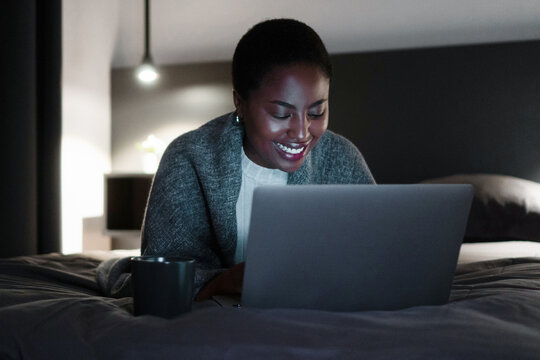 Smiling black woman, holding cup of coffee, lying on bed, looking at laptop screen