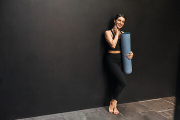 Full length young caucasian girl with slim body shape holds mat standing near wall with space for text. Brunette wears black sports top and leggings. Healthy lifestyle concept