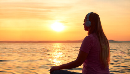 Young relaxed woman sits in headphones, watches the beautiful sunset on the sea on a summer evening