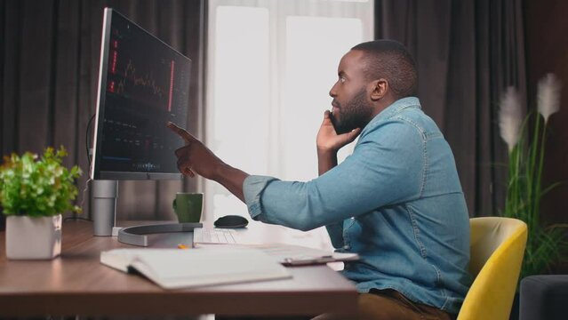 Side view portrait on african american trader, speaking on phone and pointing on screen with stock graphs. Crypto currency analytics. Graphs analysis, stock market. 4k. Office atmosphere.