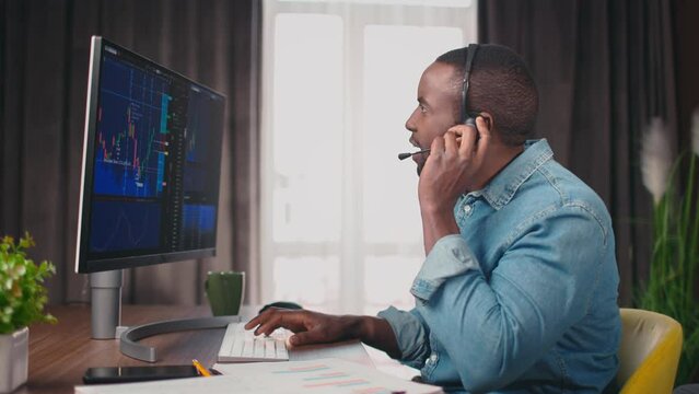 Side view portrait on african american trader, speaking in headphones and typing, looking on screen with stock graphs. Crypto currency analytics. Graphs analysis, stock market. 4k. Office atmosphere.