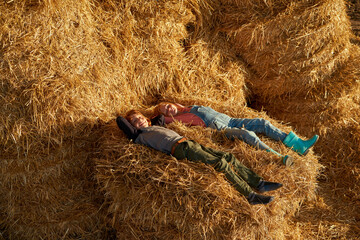 Brother and sister resting on hay bale in barn - obrazy, fototapety, plakaty