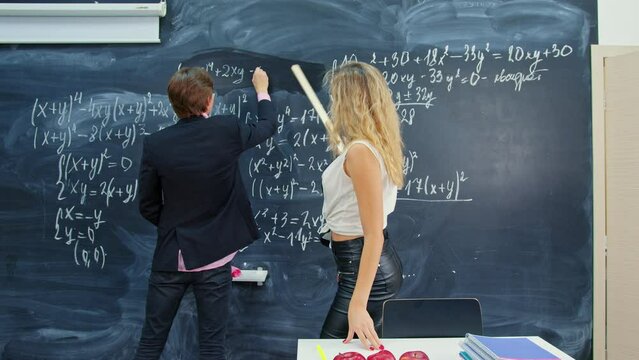 Student solves mathematical task at blackboard in classroom. Sexy young teacher woman comes closer, points out error with ruler. Provocatively dressed mathematician is in algebra lesson at college