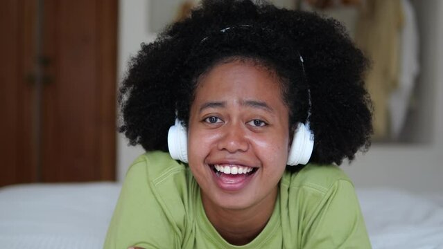 A singing woman lies on a bed in a room, listens to music on headphones and sings along with a smile. Mixing races of Africans and Asians in America. The carefree life of a young woman.
