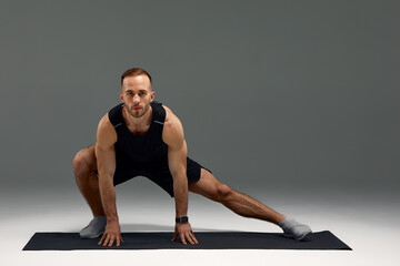 Obraz premium Attractive healthy young sports man stretching on fitness mat indoors, over gray wall background