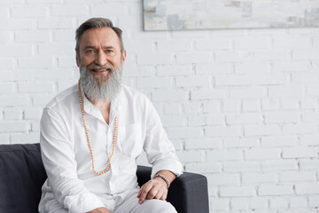 happy master guru in white shirt and beads sitting on sofa and smiling at camera. - obrazy, fototapety, plakaty