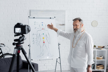 senior ayurveda coach pointing at human chakras system on flip chart in front of digital camera.