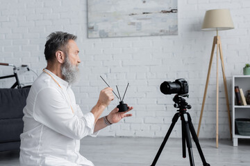 side view of ayurveda guru holding diffuser with aroma sticks near digital camera.