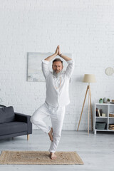 senior bearded man showing anjani mudra with raised hands while meditating in tree pose.