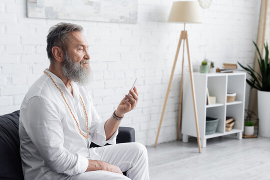 bearded guru man in white clothes sitting on sofa and looking at mobile phone.