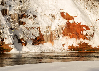 magical sunny winter day, landscape with red sandstone cliffs that are snowy with snow, frozen icicles on the cliff wall