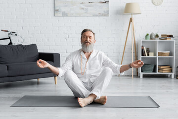 bearded guru coach meditating in easy pose with closed eyes in living room.
