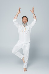 full length of senior yoga coach meditating in tree pose with raised hands on grey.