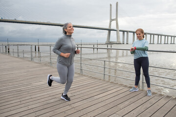 Joyful senior friends jogging on embankment. Women in sportive clothes exercising on cloudy day. Sport, friendship concept