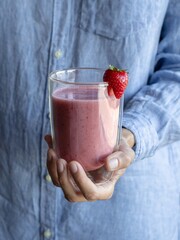 Female hand holding glass of pink smoothie. Cold strawberry-banana shake with coconut milk. Raw healthy drink with fruits and berries decorated strawberry. Vegan smoothie, blue shirt on background
