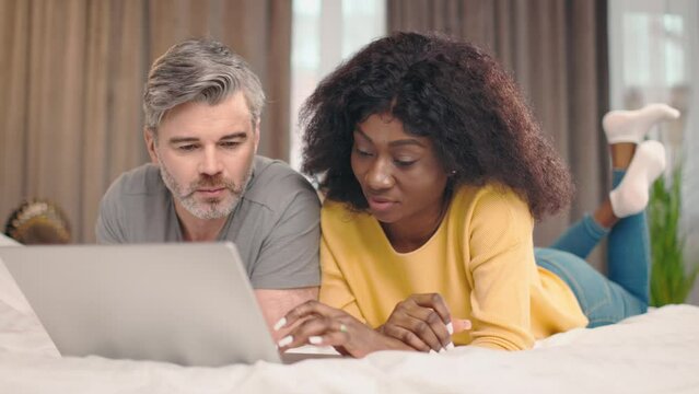 Happy middle aged man and young african american woman using laptop computer at home watching online show lying in bed together. Couple looking at gadget screen making video call chatting in bedroom