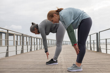 Contented friends exercising on embankment. Women in sportive clothes stretching on cloudy day....