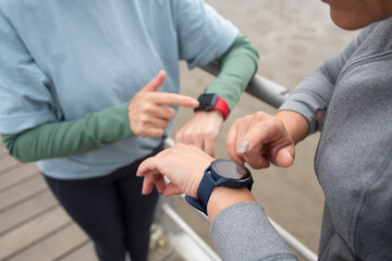 Close-up of friends checking pulse after training. Women in sportive clothes looking at wristwatch on embankment on cloudy day. Sport, friendship concept