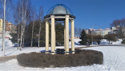 A rotunda with concrete columns stands by the walkway at the edge of a city park. Next to it are...