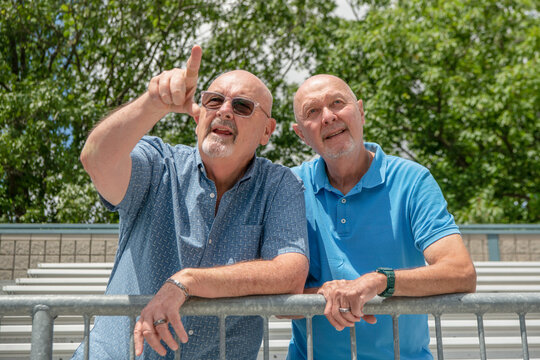 Two Men, A Mature LGBT Gay Married Couple, Lean Against A Railing In A Stadium And Are Pointing And Looking Up At Distance.