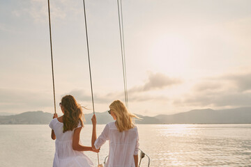 Two friends on holiday cruise together holding rigging cables enjoying the sunset. Two women enjoying the sunset together holding boat rigging cables - obrazy, fototapety, plakaty