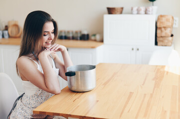 Young Woman in an Apron Sits at Wooden Table and Looks into a Saucepan. Think what to cook