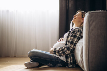 Pregnant woman sitting on floor by the coach and hugging her belly