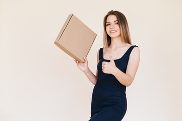Fototapeta premium Woman in Black Dress Showing Thumb Up on Beige Background, Holds Cardboard Box in her Hands