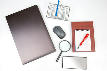 Laptop, magnifying glass, computer mouse, smartphone, pen, notepad and grocery basket on a white background. Items for business and buying goods online