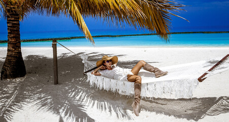 Elegant woman in fashionable dress and big hat on tropical beach on Maldives island. Summer fashion. Female model is posing on the sandy beach. Maldives landscape. Luxury Travel.