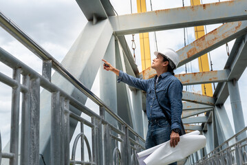Asian male engineer standing on the bridge and is examining the strength of the bridge's structure,...