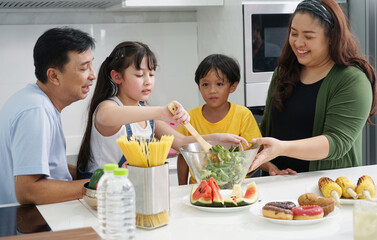 family with healthy cooking concept Father, mother and brother help each other make salad, and their eight-year-old daughter makes a salad for breakfast in the kitchen.