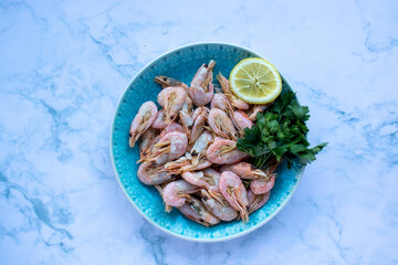 Сooked shrimp with parsley and lemon in a blue ceramic bowl on a white marble table background. Healthy Mediterranean seafood. Top view, flat lay, copy space