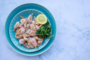 Сooked shrimp with parsley and lemon in a blue ceramic bowl on a white marble table background. Healthy Mediterranean seafood. Top view, flat lay, copy space
