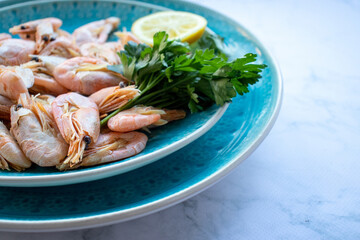Сooked shrimp with parsley and lemon in a blue ceramic bowl on a white marble table background. Healthy Mediterranean seafood. Top view, flat lay, copy space