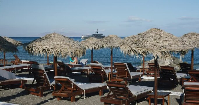 People at oceanside beach lounge relaxing under wooden umbrellas in chairs near water Relaxing chairs at beach lounge overlooking oceanside with mega yacht near in Santorini, Greece