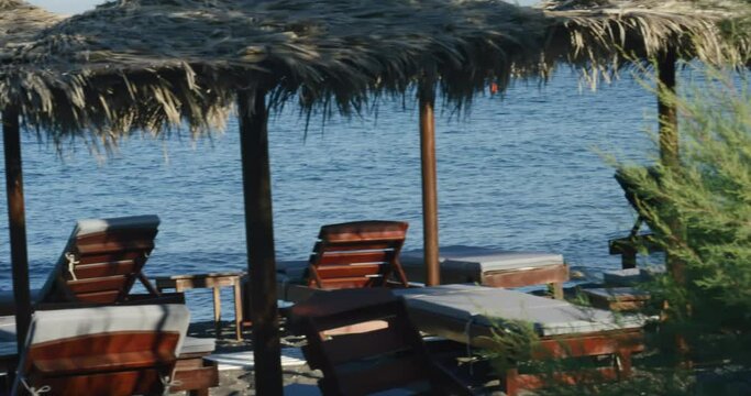 Close view of multiple relaxing lounge chairs on volcanic black sand at beach club near ocean in Santorini, Greece