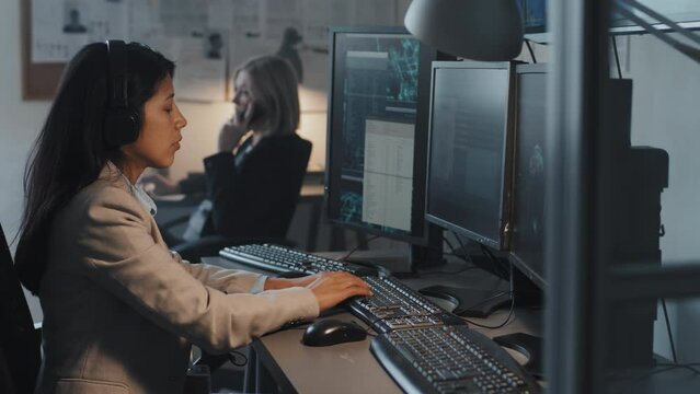 Medium side view of female Hispanic FBI agent wearing headset, using computer, sitting at desk in office at daytime