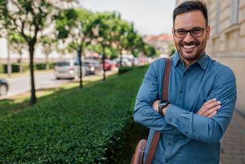 Portrait of happy businessman carrying bag standing with arms crossed in the city