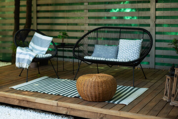 Fototapeta na wymiar Modern lounge outdoors in backyard. Terrace house with wooden floor, comfortable sofa, armchair and wicker ottoman. Cozy space in patio or balcony for relax. Wooden veranda with garden furniture. 