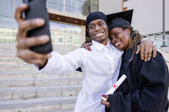 Happy son posing for selfie with college graduate mom in cap and gown