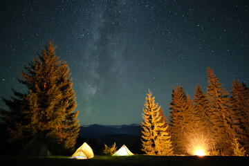 Bright illuminated tourist tents near glowing bonfire on camping site in dark mountains under night sky with sparkling stars. Active lifestyle concept