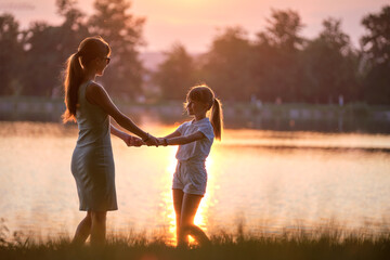 Naklejka na ściany i meble Happy mom and daughter girl relaxing holding hands enjoying time together in summer park at sunset. Family love and relationship concept