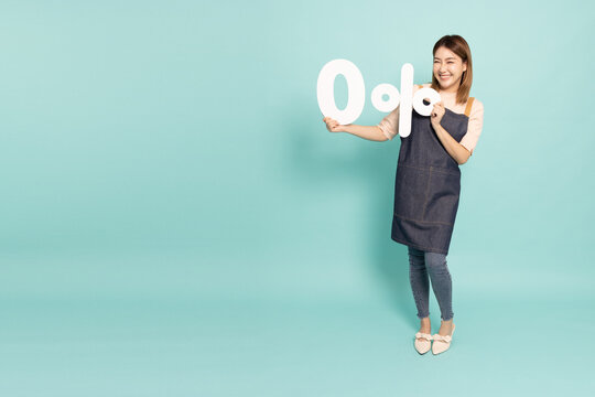 Young Asian Woman Housewife Wearing Kitchen Apron Cooking And Holding Zero Percent Or 0% Isolated On Green Background, Full Body Composition