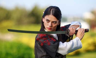 Young asian woman in traditional kimono trains in a fighting stance close-up portrait with katana sword samurai warrior girl in green summer garden