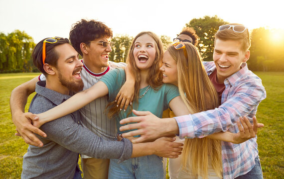 Group of funny, excited friends fooling around and having fun in the park. Bunch of happy, joyful, smiling people standing on a green lawn and all together hugging a cheerful beautiful young girl