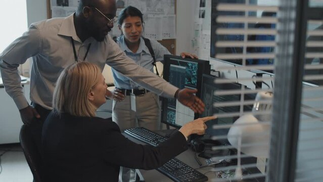 Tilting up slow motion of three multiethnic detectives at desk in office at daytime, searching digital database, talking, using computer
