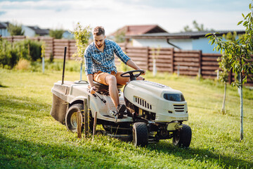 Portrait of Industrial gardener driving a riding lawn mower in in a garden. Professional landscaper using tractor and mowing lawn. - obrazy, fototapety, plakaty