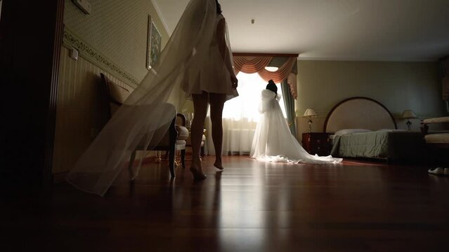 Bride enters the room in long bridal veil, strokes and touches her wedding dress with her hands. Woman admires her white dress.